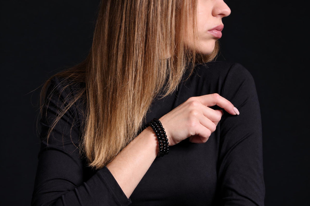 A woman with long hair wears a black beaded bracelet on her wrist against a dark background.