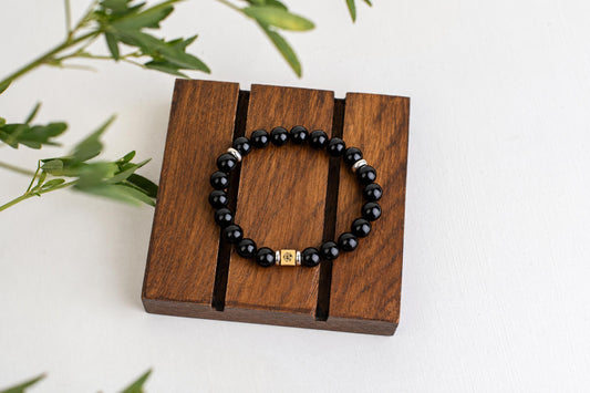 A black beaded bracelet with silver and gold-toned accents rests on a dark wooden stand against a white surface.