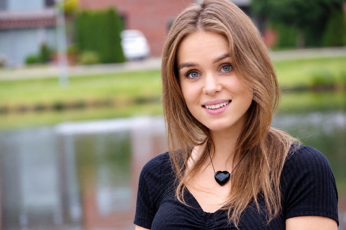 A woman wears a black heart-shaped pendant on a thin cord against a dark top, outdoors near water.