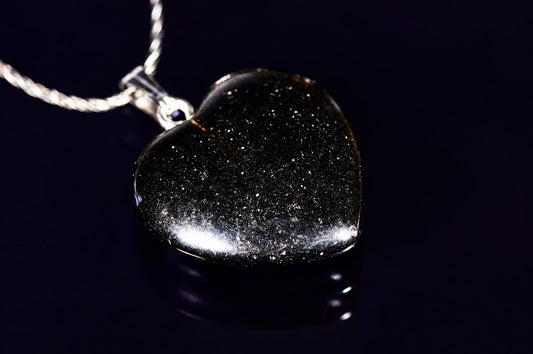A heart-shaped pendant with a black, speckled surface hangs from a silver-toned chain on a dark reflective surface.