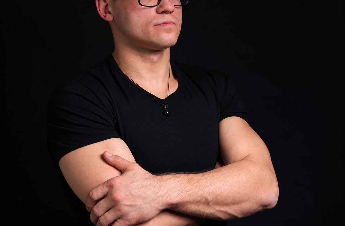 A man in a black t-shirt wears a necklace with a small, round, dark pendant on a thin cord against a dark background.