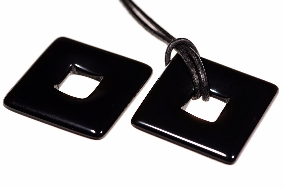Two glossy black square pendants with rounded corners and a central square cutout, one attached to a dark cord on a white surface.