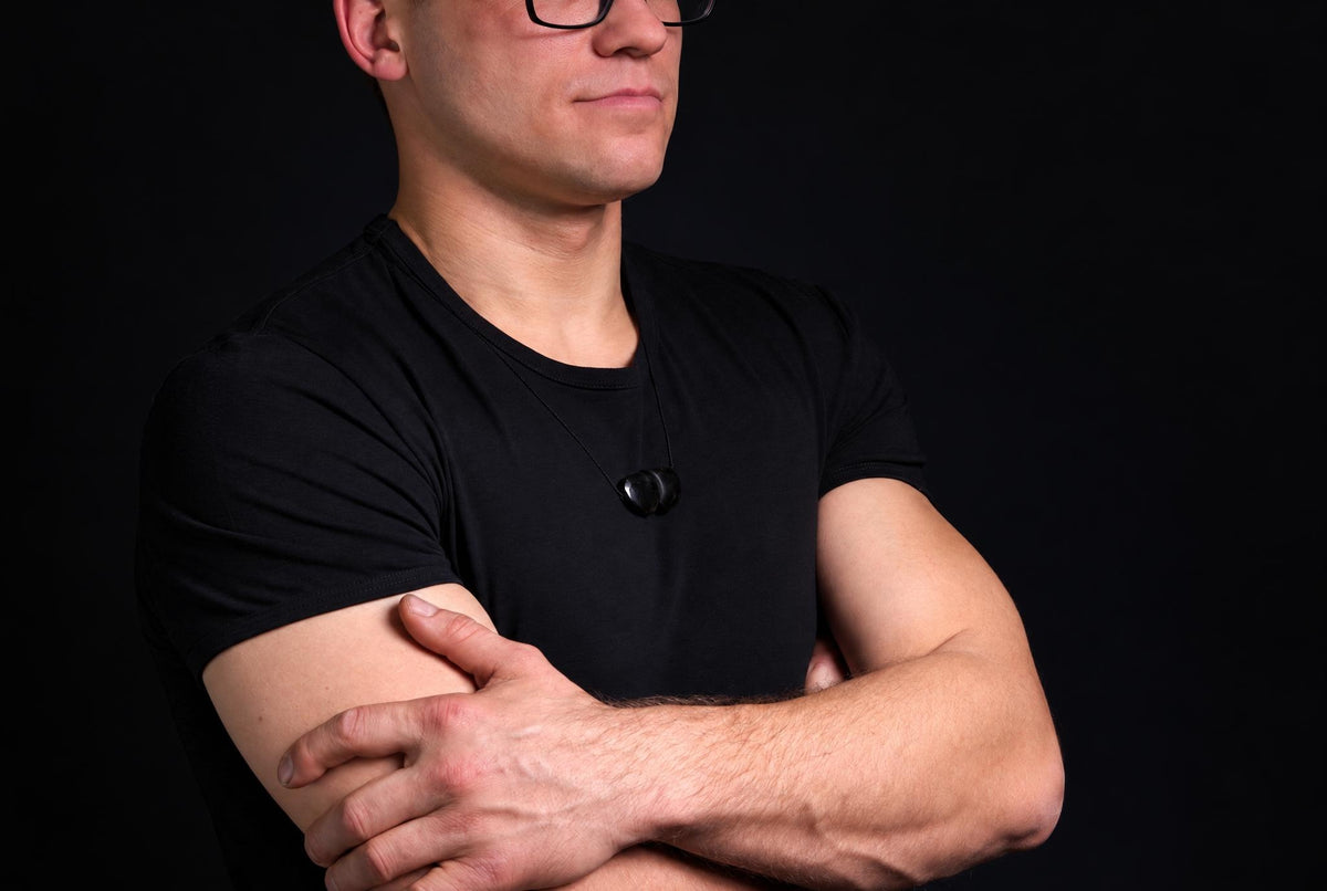 A man in a black t-shirt wears a necklace with two dark, smooth stones on a thin cord against a dark background.