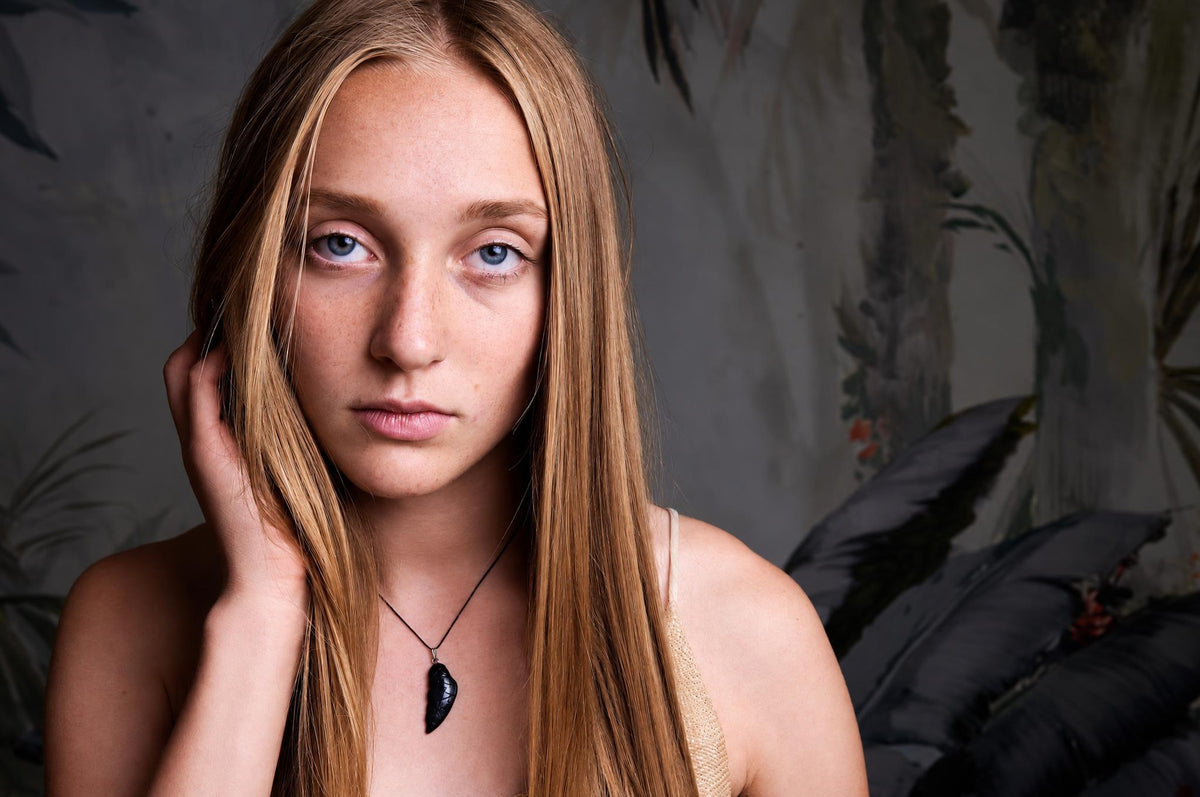 A woman with long blonde hair wears a black pendant shaped like a wing on a thin cord against a dark surface.