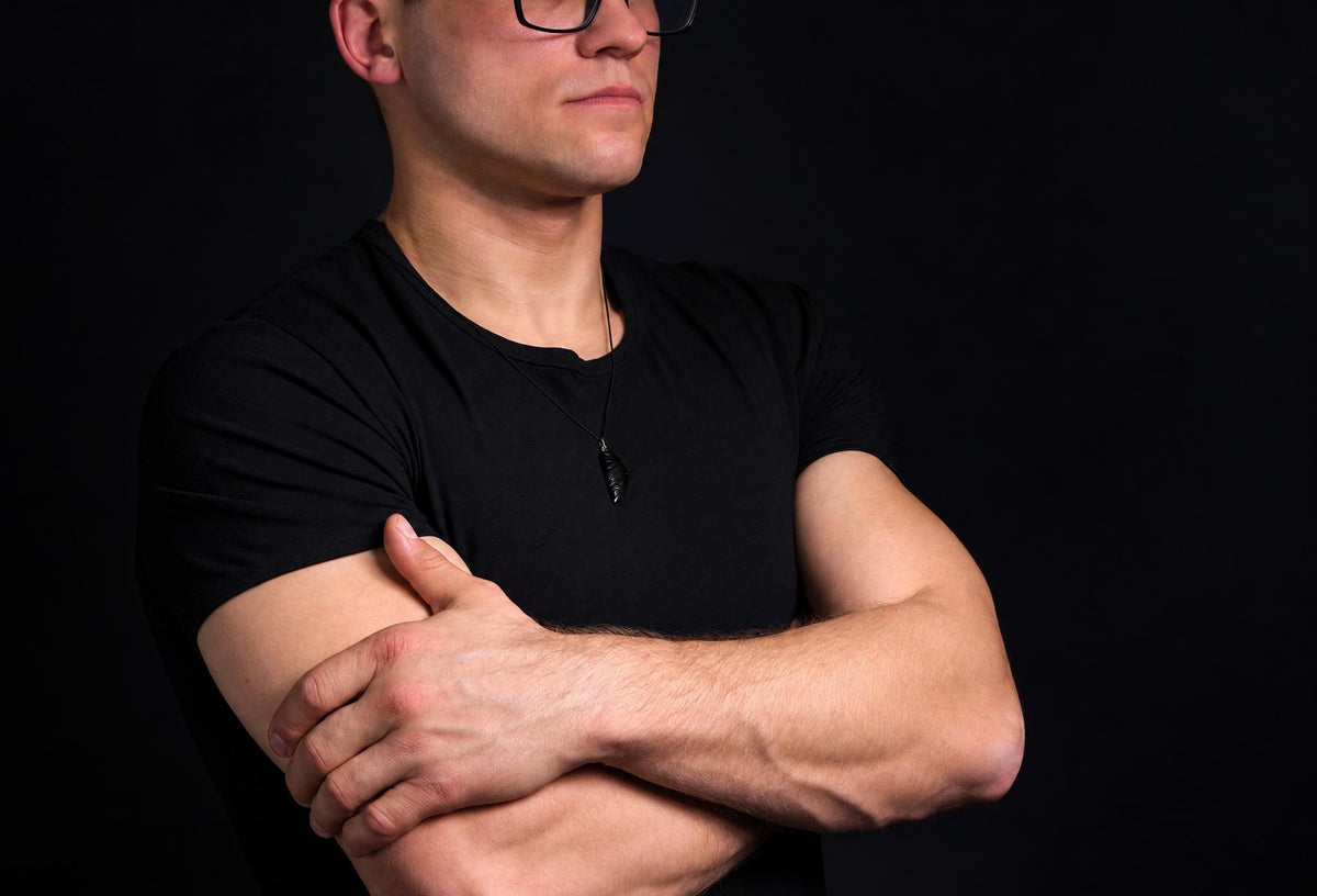 A man in a black t-shirt wears a dark pendant shaped like a wing on a thin cord against a dark background.