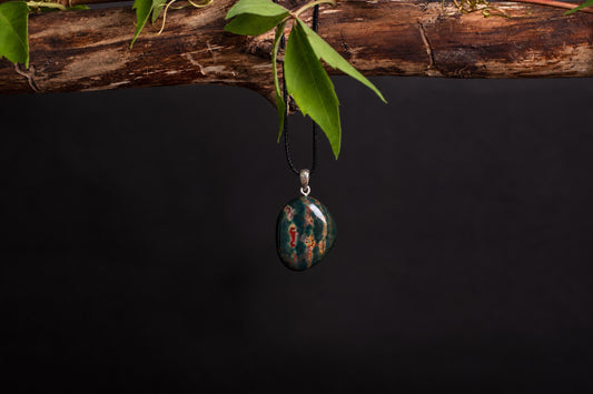 A polished oval pendant with green and red patterns hangs from a black cord on a dark surface.