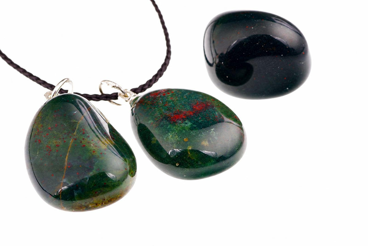 Three polished green stones with red speckles, two on dark cords and one loose, against a white background.