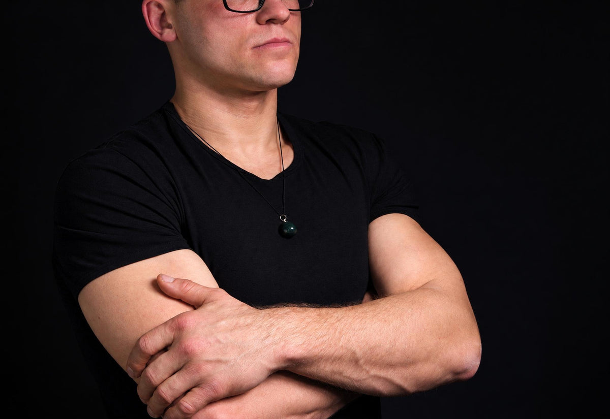 A man wears a black t-shirt and glasses, with arms crossed against a dark background. He has a necklace with a round, dark green pendant.