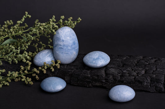 Five smooth, pale blue stones rest on a dark wooden surface beside green sprigs against a black background.