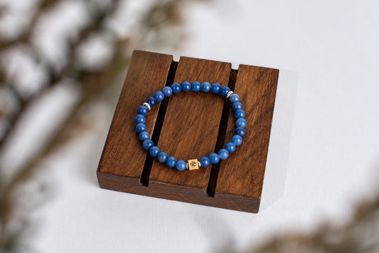 A blue beaded bracelet with a gold-toned square charm rests on a dark wooden display stand.