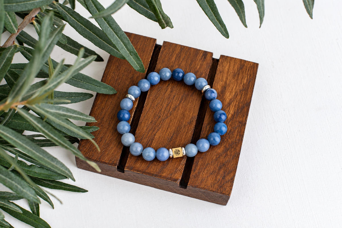 A blue quartz beaded bracelet with silver-toned spacers and a gold-coloured square charm rests on a dark wooden surface beside green leaves.