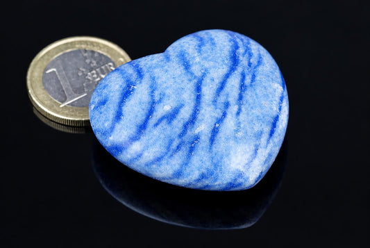 A polished blue quartz heart with white streaks rests on a dark surface next to a one-euro coin.