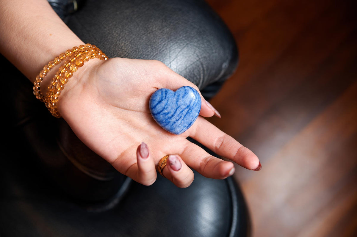 A hand holds a polished, blue heart-shaped stone with lighter swirls on a dark surface.