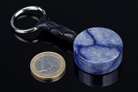 A round blue stone keychain with a braided black leather strap and metal ring, next to a one-euro coin on a dark surface.