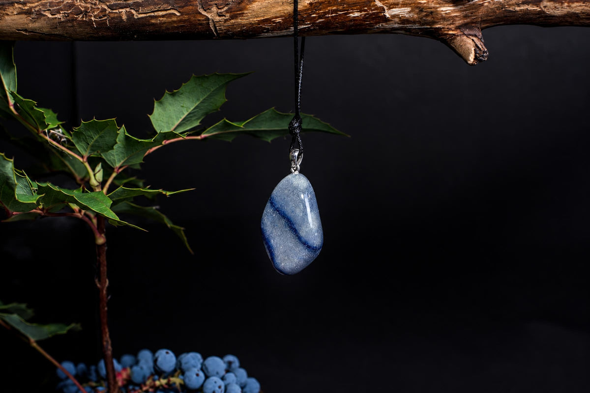 A polished blue quartz pendant with a silver bail hangs from a black cord against a dark background, near green leaves.