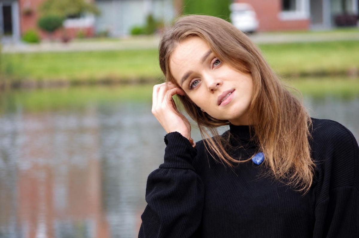 A woman wears a black turtleneck sweater with a blue pendant necklace against a blurred outdoor background.