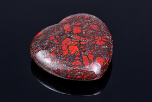 A polished heart-shaped stone with a dark brown base and bright red, jagged patterns on a dark reflective surface.