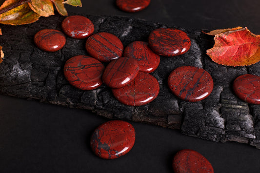 Polished red palmstones with black veins rest on a dark, cracked wooden surface, surrounded by autumn leaves.