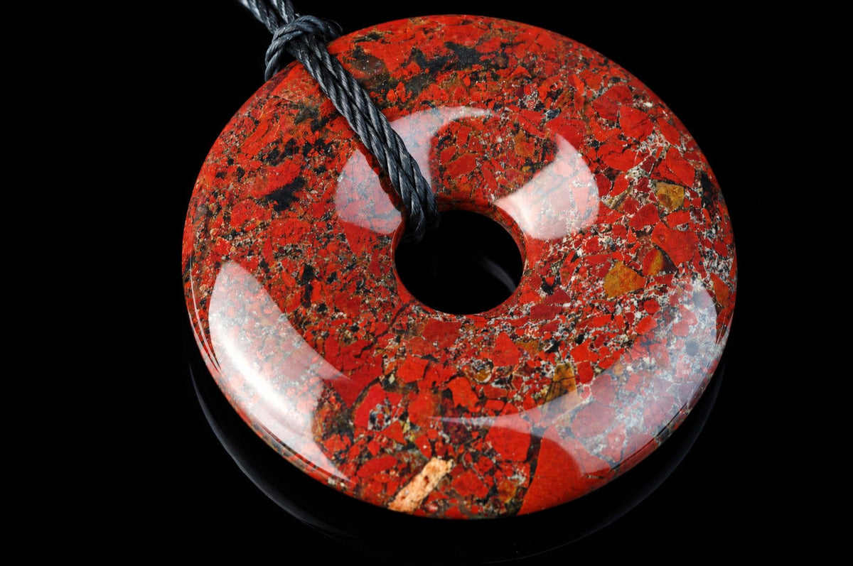 A polished, red and black speckled donut-shaped pendant with a central hole, threaded on a dark cord against a black background.