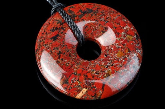 A polished, red and black speckled donut-shaped pendant with a central hole, threaded on a dark cord against a black background.