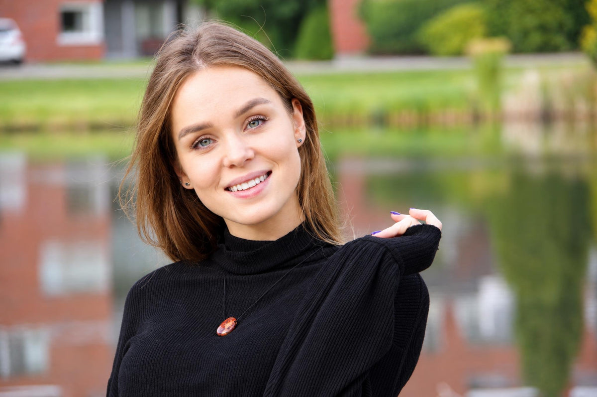 A woman wears a black turtleneck with an oval, reddish-brown pendant on a thin cord against a blurred outdoor background.