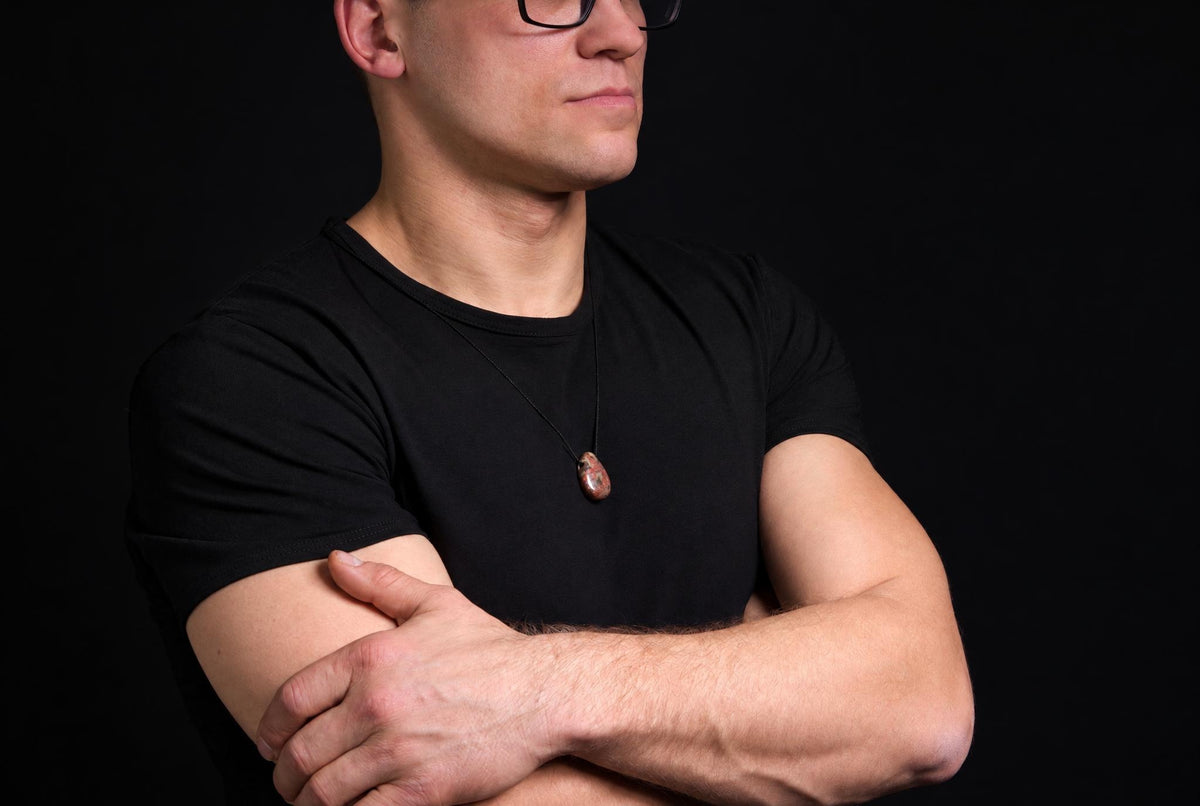 A man in a black t-shirt wears a necklace with an oval pendant of reddish-brown and white speckled stone against a dark background.