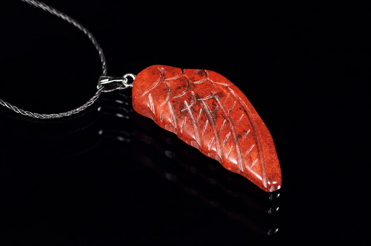 A red, carved pendant shaped like a wing with incised lines, attached to a silver-toned chain on a dark reflective surface.