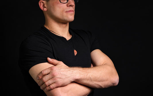 A man in a black t-shirt wears a pendant with an irregular, reddish-brown stone on a dark cord against a black background.