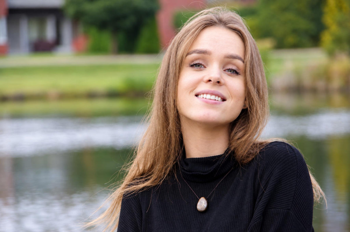 A woman with long light brown hair smiles while wearing a black turtleneck and a pendant necklace with a teardrop-shaped stone on a dark surface.