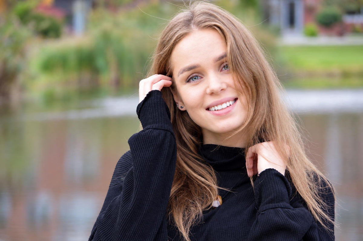 A smiling woman with long brown hair wearing a black turtleneck sweater, posing outdoors near water.