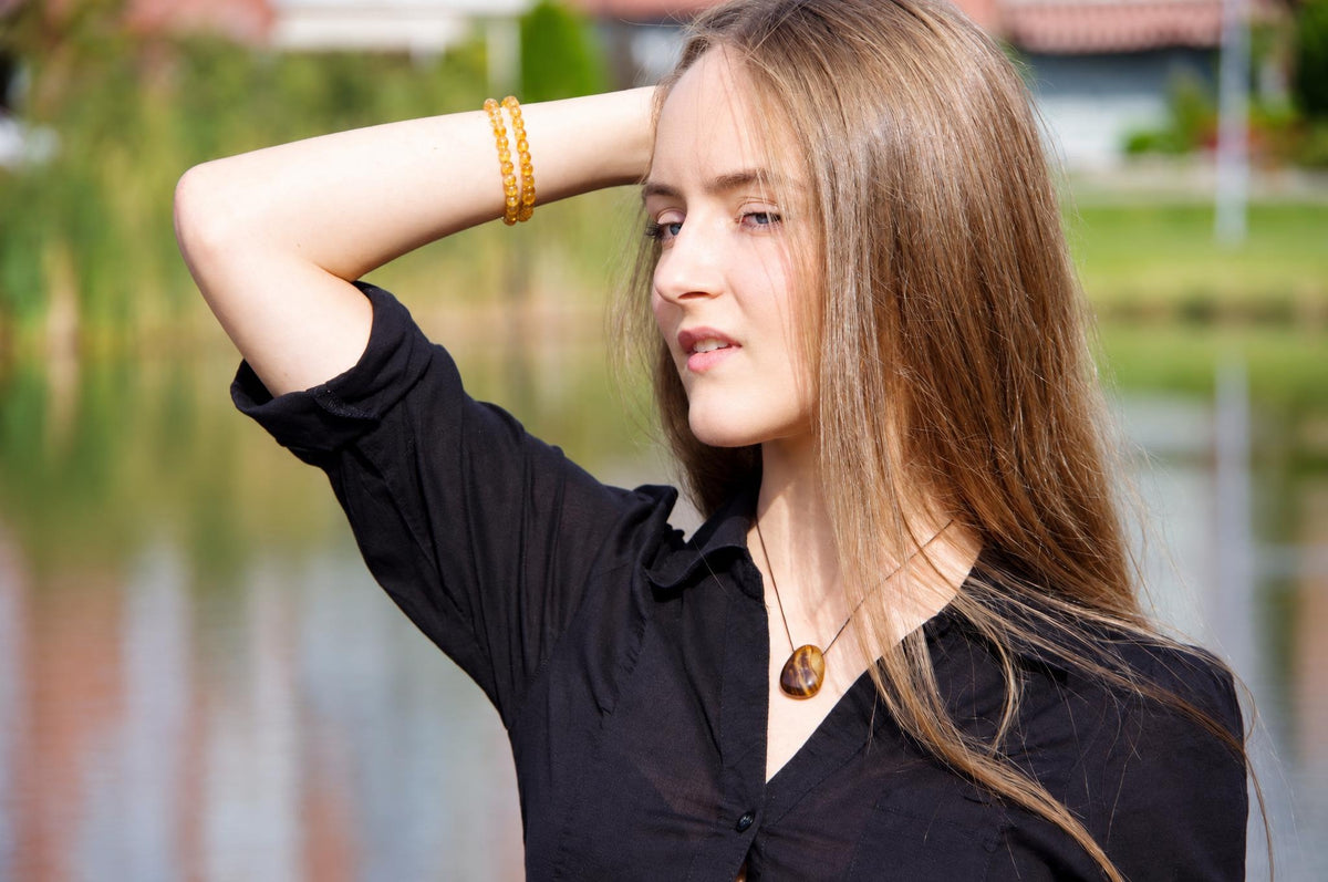 A woman in a black shirt wears a brown teardrop pendant outdoors, with one arm raised and blurred greenery behind her.
