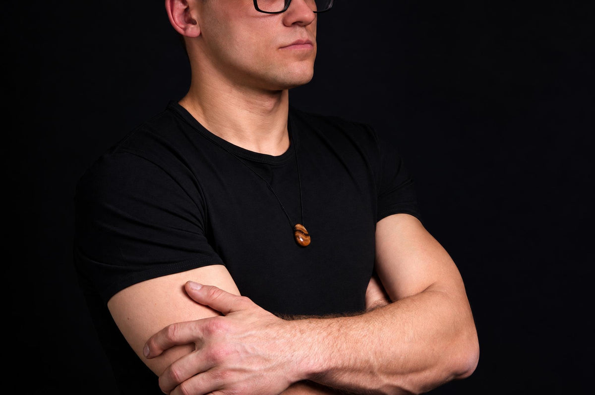 A man in a black t-shirt wears a brown pendant on a dark surface against a black background.