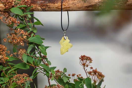 A yellow-green, irregularly shaped pendant hangs from a black cord on a wooden branch, surrounded by green leaves and brown flowers.