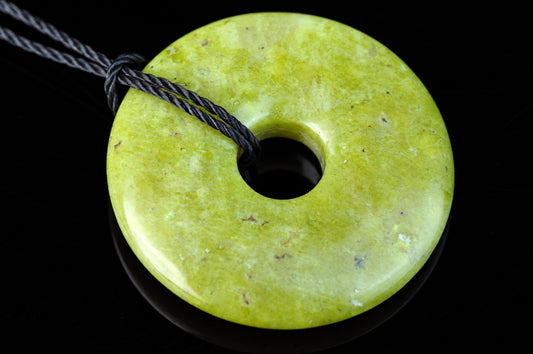 A round, pale green pendant with a central hole and a dark cord looped through it, resting on a black surface.