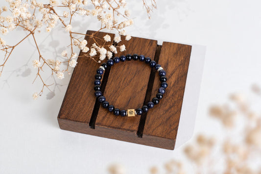 A dark blue beaded bracelet with a gold-toned square charm rests on a wooden display stand, surrounded by dried flowers.