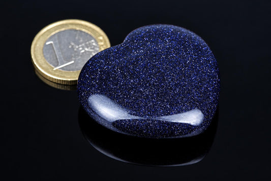 A polished deep-blue heart with sparkling flecks rests beside a coin on a black reflective surface.