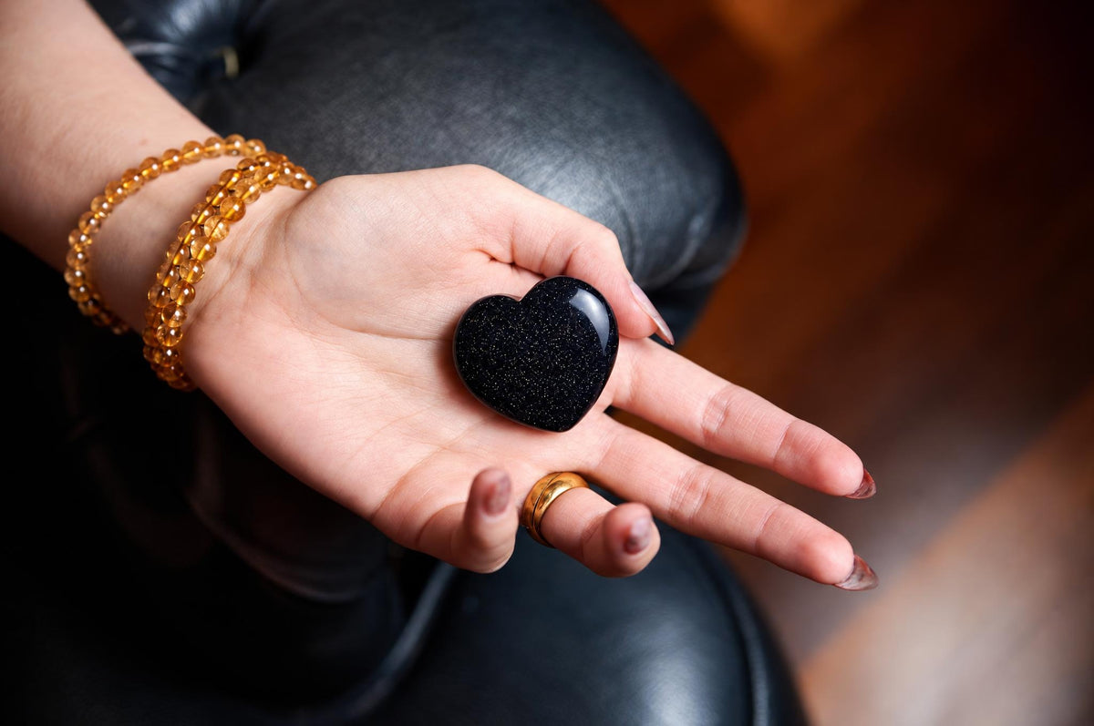 A hand holds a polished, black heart-shaped stone with a glittery texture against a dark leather surface.