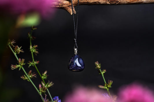 A dark blue, teardrop-shaped pendant with a white crescent moon hangs from a black cord on a wooden branch against a dark background.
