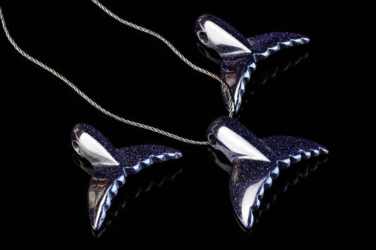 Dolphin-tail pendants with dark blue sparkling surfaces and black cords lie on a black reflective background.