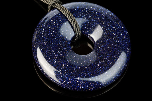 A round, donut-shaped pendant with a dark blue glittery surface and a central hole, suspended by a grey cord against a black background.