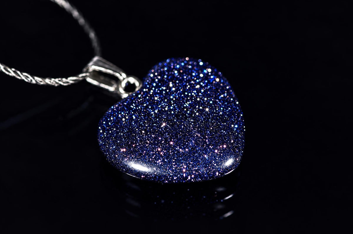 A heart-shaped pendant with blue glitter on a dark surface, attached to a silver-toned chain.
