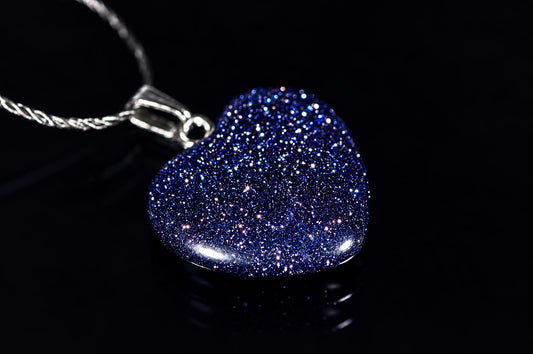 A heart-shaped pendant with blue glitter on a dark surface, attached to a silver-toned chain.