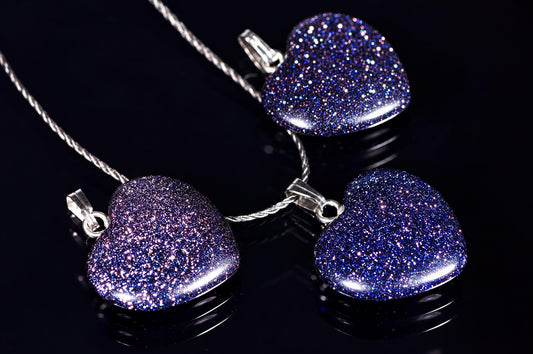 Three heart-shaped pendants with glittery blue and purple surfaces hang from silver-toned chains on a dark, reflective surface.