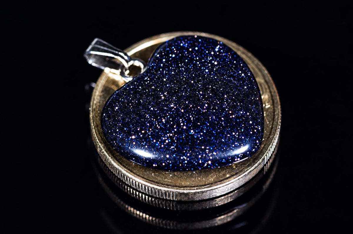 A heart-shaped pendant with a dark blue, glittering stone set in a gold-toned bezel on a black reflective surface.