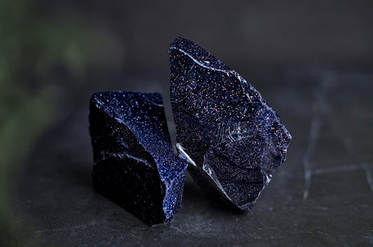 Two dark blue, glittering raw stone fragments rest on a dark surface.