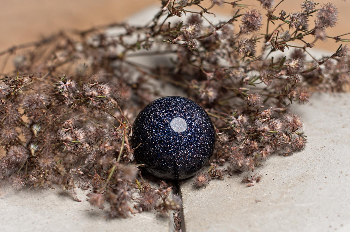 A dark blue, glittery sphere with a white spot rests on a pale surface among dried brown flowers.