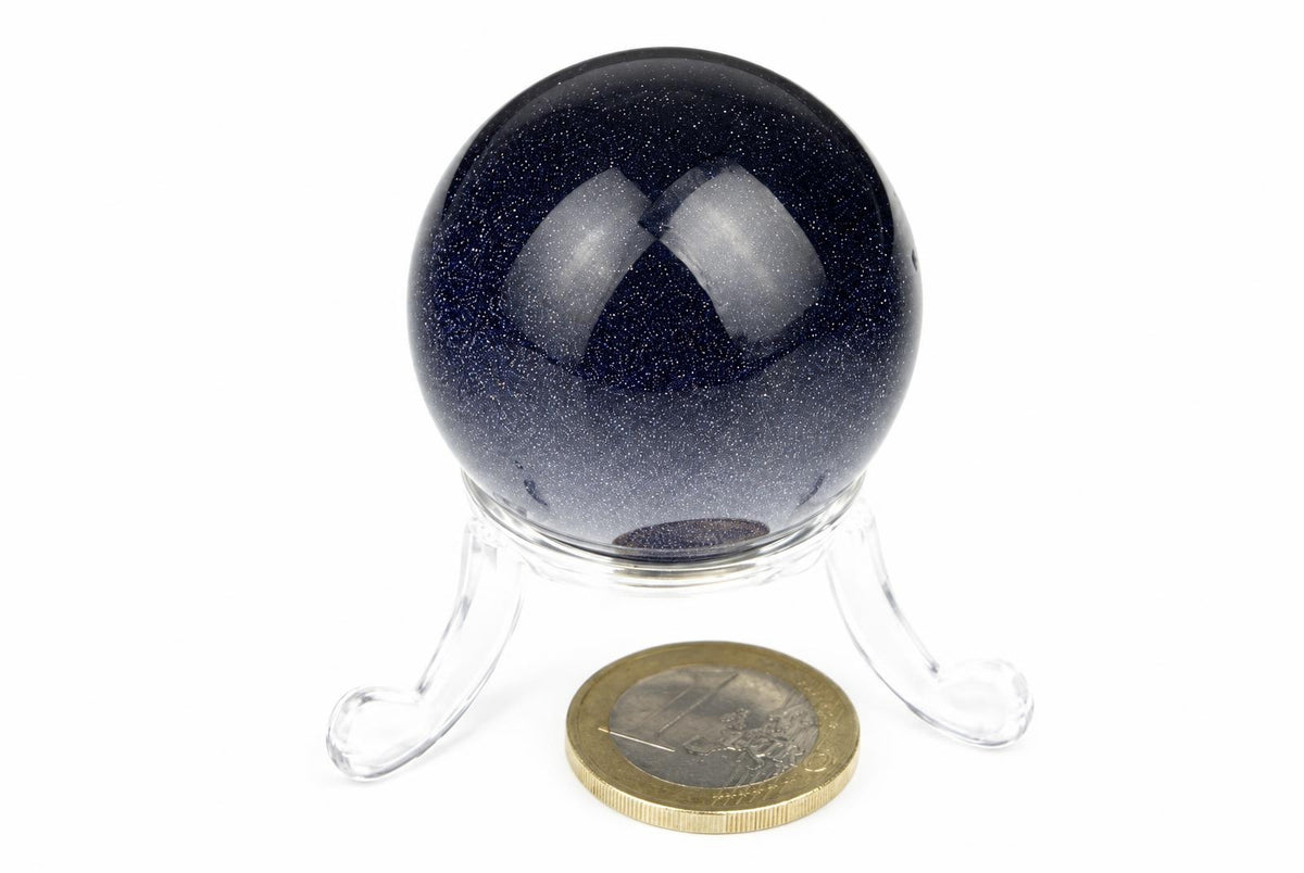 A polished dark blue sphere with sparkling flecks sits on a clear stand above a coin on a white surface.