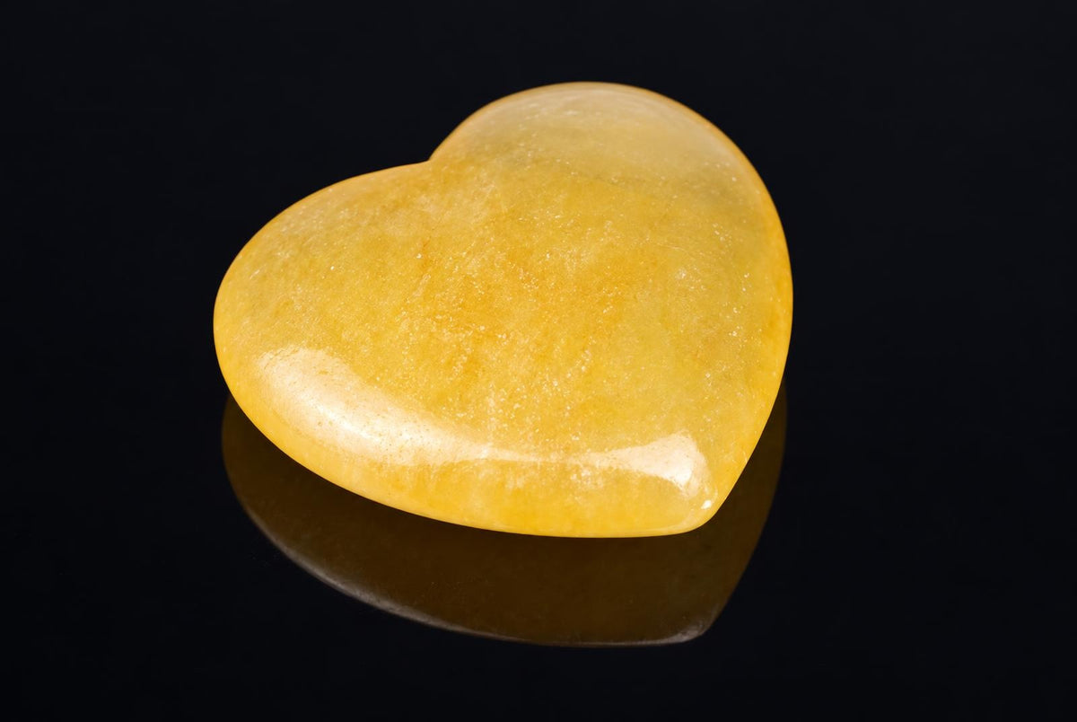A polished, heart-shaped Calcite stone with a smooth yellow surface rests on a dark reflective surface.