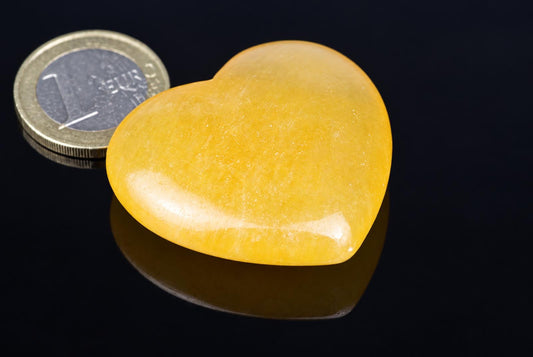 A polished, yellow heart-shaped Calcite stone rests on a dark surface next to a Euro coin.