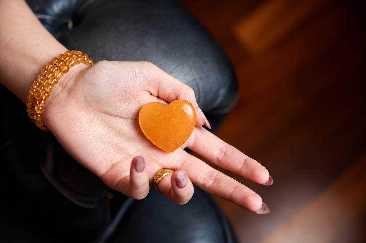 A hand holds a smooth, orange heart-shaped stone on a dark surface.
